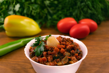 Stew of finely chopped stewed vegetables on a background of whole ripe tomatoes, peppers, parsley leaves and dill on a wooden surface.