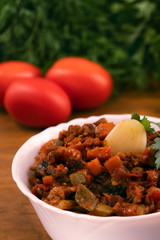 Braised eggplant with courgettes, peppers, carrots and tomatoes in a deep white bowl on a wooden surface against a background of parsley and dill leaves.
