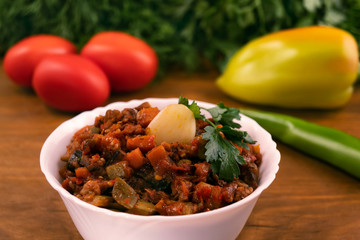 Stew of stewed vegetables in a white plate with a clove of garlic on the background of whole tomatoes, pepper and parsley leaves and dill.