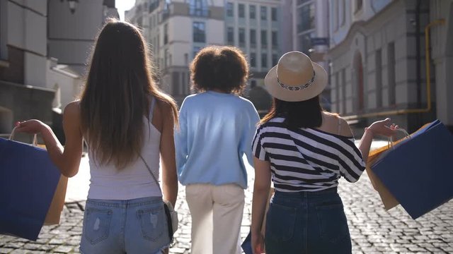 Back view of three multi ethnic female friends walking along shopping street in city in summer and putting paper shopping bags on their shoulders. Asian, mixed race and caucasian girls shopping