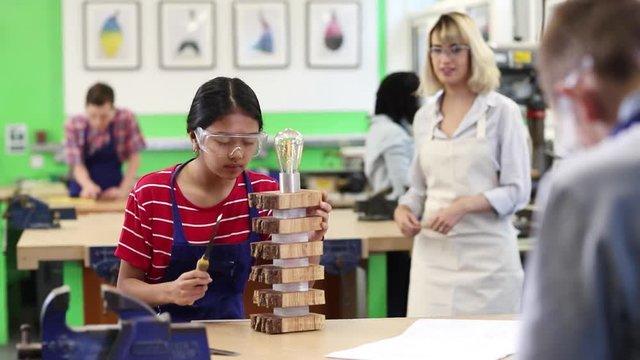 Teacher Helping Female High School Student Building Lamp In Woodwork Lesson