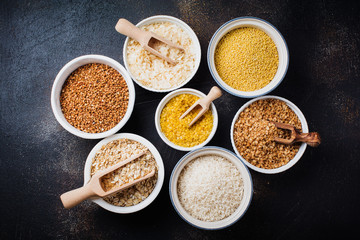 Variety of cereal flakes rice, millet, buckwheat, oatmeal. Superfood in white ceramic bowls on dark old   concrete background. Top view.