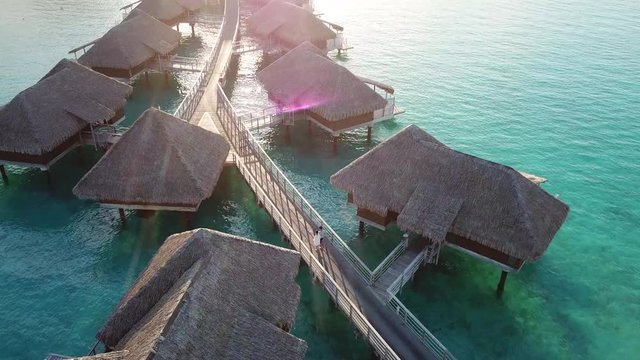 A couple walking down the wooden boardwalks that stretch into the Pacific Ocean, passing through dreamy overwater bungalows with beams of setting sun shedding on them in Bora Bora