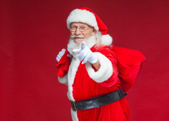 Christmas. Smiling Santa Claus in white gloves with a bag of gifts behind him points his index finger into the camera. Isolated on red background.
