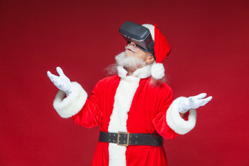 Christmas. Santa Claus in black virtual reality glasses makes gestures with his hands. Surprise, emotion. New technology. Isolated on red background.