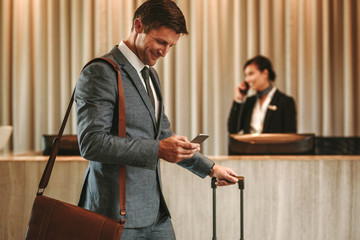 Businessman in hotel hallway with cellphone and luggage - obrazy, fototapety, plakaty