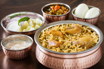 Chicken biryani with traditional sides - Closeup view from the top of delicious Indian chicken biryani served in authentic copper utensils with salad (raita), gravy and egg. Natural light used.