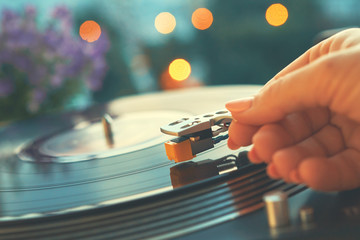 Vinyl player on a sunset background with a bokeh lights of the evening city. The girl's hand places the needle on a rotating black vinyl plate. Included gramophone and rotating plate