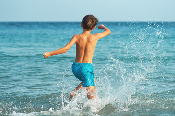 A happy little boy is happily running and splashing in the sea water in summer on a warm, sunny day. Large splashes of water close-up. A child is playing on the seashore