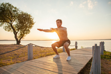 Fitness man training air squat exercise on beach outside. Fit male exercising crossfit outside. Young handsome caucasian male fitness model and instructor outdoors.