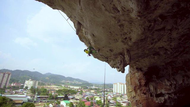 A man rock climbs out of a cave and clips the rope into a quickdraw as he tries hard.