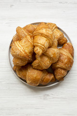 Fresh golden croissants on grey round plate on white wooden surface, overhead view. From above, top view.