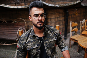 Fototapeta na wymiar Close up portrait of awesome beautiful tall ararbian beard macho man in glasses and military jacket posed outdoor, sitting on chair against wooden house.