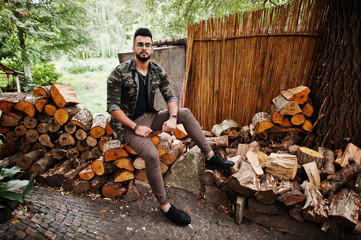 Awesome beautiful tall ararbian beard macho man in glasses and military jacket posed outdoor, sitting on stumps.