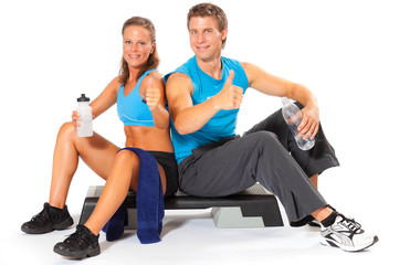 Fototapeta na wymiar Smiling man and woman sitting on single aerobic stepper holding water bottle showing thumbs up sign wearing sports wear isolated against white background