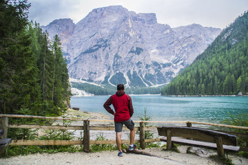 Fototapeta na wymiar View of young tourist hiker man overlooking beautiful alpine lake Lago Di Braies (Pragser wildsee) in Trentino, Dolomites mountains, Italy. Tourist popular summer attraction/destination in Europe