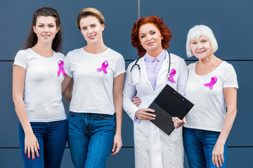 women and doctor with pink ribbons standing together and smiling at camera, breast cancer awareness...
