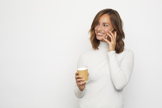 happy young woman with a cup of coffee, having a conversation on the phone looks left at copy space