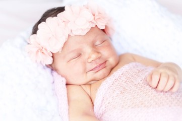 Cute Caucasian newborn baby girl with a pink flower hair band and covered by light pink blanket smiling sweetly in her sleep. Sleeping smiling infant concept image.