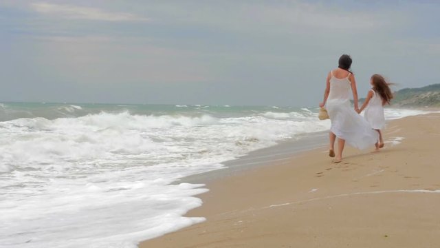 Mom and his beloved daughter are walking along the seashore, holding beach hats and chatting
