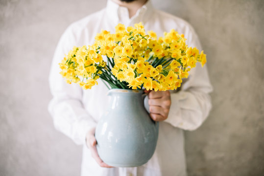 Fototapeta Very nice young man in a white shirt holding blossoming yellow Narcissus flower bouquet in a blue ceramic vase on the grey wall background