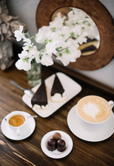 Delicious fresh morning two cups of coffee -espresso and cappuccino- white flowers in a vase, two pieces of chocolate cake and some chocolate on the white saucer on the wooden table background