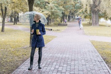 A woman of 30-40 years in a blue raincoat under a transparent umbrella walks in the park and drinks coffee.