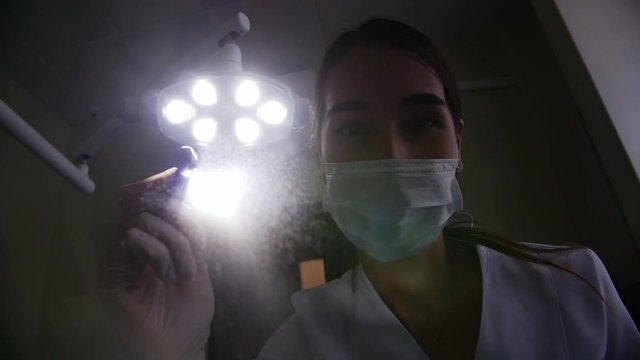 Woman dentist straightens the lamp and looks into the camera with a tool in his hand.