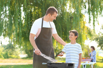 Naklejka na ściany i meble Little boy with his father cooking tasty food on barbecue grill outdoors