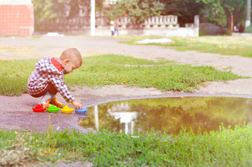 A cute baby boy plays colored boats in a puddle in yellow rubber boots and a checkered raincoat.