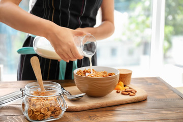 Woman pouring milk into bowl with corn flakes at table