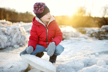 Fototapeta na wymiar Happy little girl playing with ice blocks by frozen river during an ice break. Child having fun in winter.