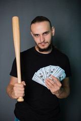 A young handsome guy in a black T-shirt on a gray background holds dollars in his hands. A man with money and a painless bat