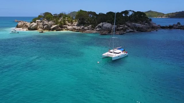 Paning aerial drone shot of a catamaran anchoring in the crystal blue water of the Seychelles in frront of tropical Cocos Islands.