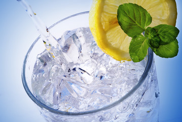 pouring fresh water into a glass with ice cubes, lemon slice and mint, light blue background