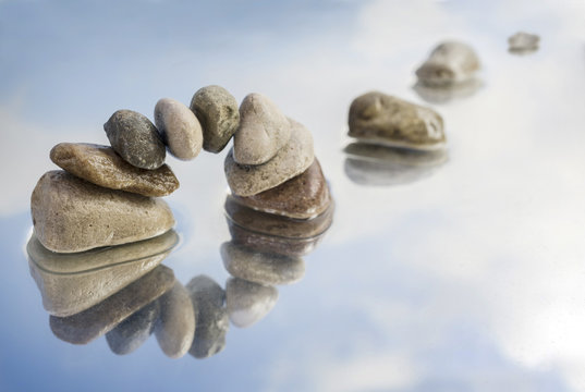 arch of balanced pebbles and stepping stones in the water with reflection, light blue sky with clouds, copy space