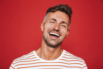 Handsome guy with stubble in striped t-shirt laughing on camera, isolated over red background