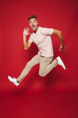 Full length photo of joyful man in striped t-shirt jumping and screaming, isolated over red background