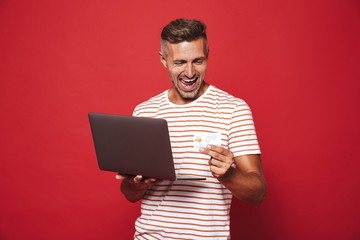 Image of bearded man in striped t-shirt smiling while holding credit card and laptop, isolated over...