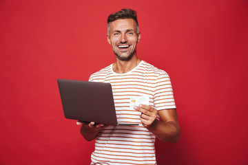 Image of brunette man in striped t-shirt smiling while holding credit card and laptop isolated over...