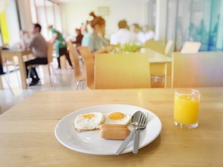 Homemade delicious american breakfast with sunny side up fried egg and sausages in white dish serve with orange juice on the wooden table with background of people at restaurant, hotel in the morning