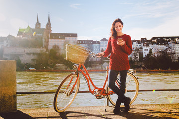 theme tourism on bicycle and modern technology. Beautiful young caucasian woman stands near red retro bicycle on riverside river Rhine Basel Swiss winter warm sunny weather uses the phone in hand.