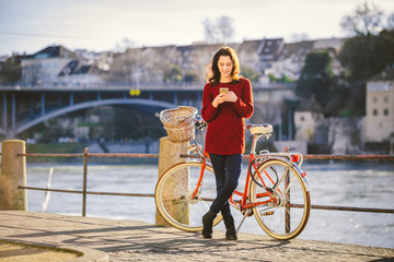 theme tourism on bicycle and modern technology. Beautiful young caucasian woman stands near red retro bicycle on riverside river Rhine Basel Swiss winter warm sunny weather uses the phone in hand.