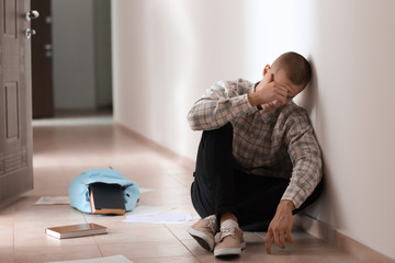 Sad teenage boy sitting on floor indoors. Bullying at school