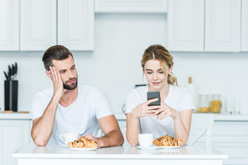 bored man looking at smiling girlfriend using smartphone during breakfast