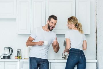 cheerful young couple laughing together in kitchen, man showing thumb up and smiling at camera
