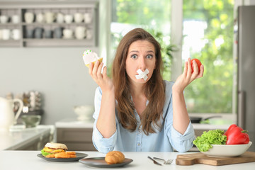 Stressful woman with taped mouth and different products in kitchen. Choice between healthy and...