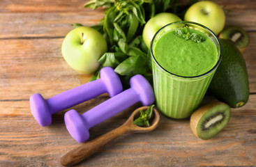 Glass of tasty green smoothie with ingredients and dumbbells on wooden table. Diet concept