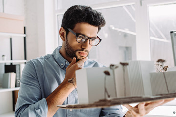 Fototapeta premium pensive architect in glasses touching chin and looking at architecture model in office