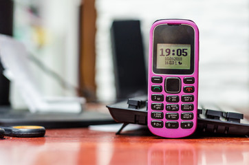 Old button mobile phone pink color on the desk near a computer keyboard. Clock and indicators on the black and white screen.